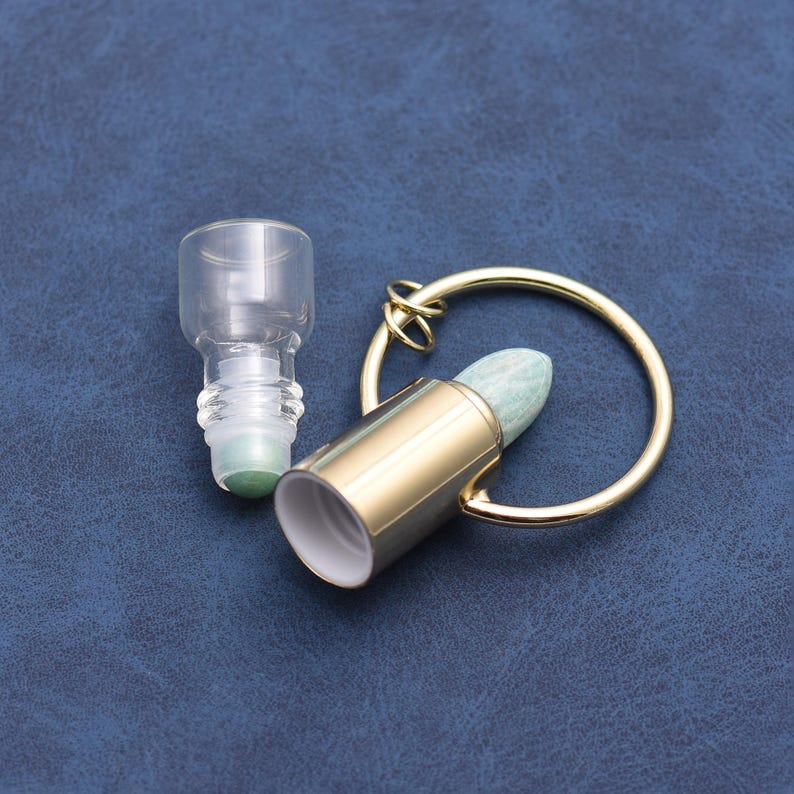 May include: A clear glass roller bottle and a gold-colored metal rollerball with a light green stone tip. The rollerball is attached to a gold-colored ring. The items are on a dark blue surface.
