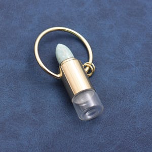 May include: A gold-colored metal essential oil bottle with a clear glass base and a light green stone top. The bottle has a gold ring attached for carrying. The background is a dark blue.