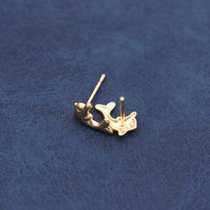 May include: A pair of gold-colored dolphin-shaped stud earrings. One earring is lying on its side, showing the post and the dolphin's body. The other earring is upright, displaying the post and the dolphin's tail and body.