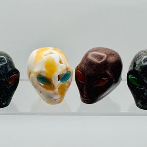 May include: Four alien head sculptures in various colors. One is black with red eyes, another is white and yellow with blue eyes, a third is brown with red eyes, and the last is black with green eyes.