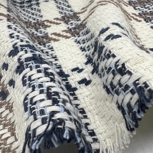 May include: Close-up of a woven fabric with a plaid pattern. The fabric features a cream-colored base with woven sections of navy blue, light blue, and brown. The edges of the fabric are frayed.