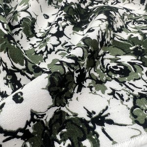以下が含まれることがあります： オリーブグリーン、黒、白の色合いで密な花柄が描かれた織物。このテキスタイルは、繰り返される植物のデザインが特徴で、糸の質感が目立ち、下端にはほつれた未仕上げの縁取りが見られます。