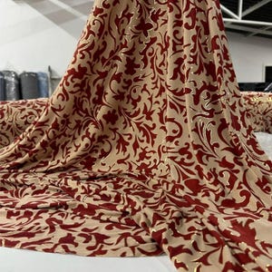 Op de afbeelding: Een beige stof met een herhalend bordeauxrood bloemmotief, mogelijk voor bekleding of gordijnen. De stof heeft een zachte textuur en gouden accenten. Het materiaal is gedrapeerd, wat de vloeiende kwaliteit laat zien. Het patroon is een klassiek ontwerp.