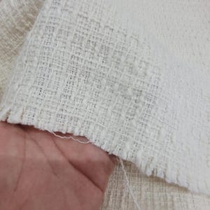 Puede incluir: Tejido blanco roto con una superficie texturizada. El tejido tiene un borde ligeramente deshilachado, revelando el tejido. El material parece ser de peso medio, adecuado para varios proyectos textiles.