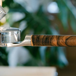 Puede incluir: Primer plano de un portafiltro de máquina de espresso cromado con un mango de madera. El mango presenta un patrón de vetas de madera marrón oscuro y negro. La máquina es parcialmente visible, con un fondo verde borroso.