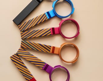 Peut inclure: Un ensemble de six poignées circulaires de type porte-filtre disposées en éventail. Chaque poignée présente une couleur métallique distincte, notamment noir, bleu, rose, orange, violet et rouge, avec un motif à rayures diagonales bleu et or.