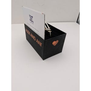 Pode incluir: Uma pulseira de metal prateado repousa dentro de uma caixa retangular preta. A caixa apresenta o texto ALEX AND ANI em letras cor de cobre, um &iacute;cone de cora&ccedil;&atilde;o cor de cobre com um padr&atilde;o de bandeira e um cart&atilde;o branco com STOMP OUT BULLYING.