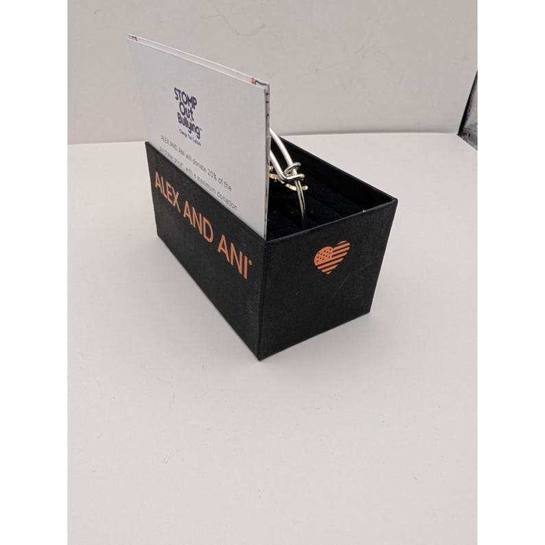 Pode incluir: Uma pulseira de metal prateado repousa dentro de uma caixa retangular preta. A caixa apresenta o texto ALEX AND ANI em letras cor de cobre, um &iacute;cone de cora&ccedil;&atilde;o cor de cobre com um padr&atilde;o de bandeira e um cart&atilde;o branco com STOMP OUT BULLYING.