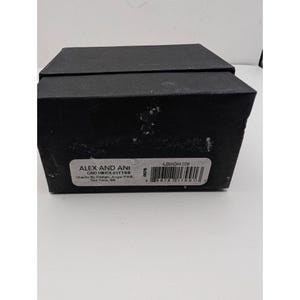 Pode incluir: Uma caixa de joias retangular preta com tampa. Uma etiqueta branca na frente exibe o texto ALEX AND ANI, CBD19HOL01TTSS, Charity By Design, Angel EWB, Two Tone, SS, ALEXANDANI.COM, e um c&oacute;digo de barras com o n&uacute;mero 8 86787 17991 3.