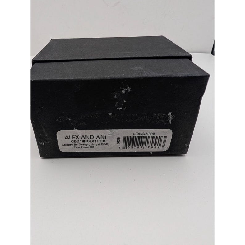 Pode incluir: Uma caixa de joias retangular preta com tampa. Uma etiqueta branca na frente exibe o texto ALEX AND ANI, CBD19HOL01TTSS, Charity By Design, Angel EWB, Two Tone, SS, ALEXANDANI.COM, e um c&oacute;digo de barras com o n&uacute;mero 8 86787 17991 3.