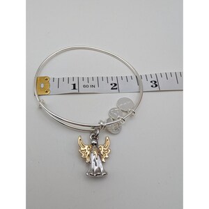 Pode incluir: Uma pulseira de fio expans&iacute;vel em tom prateado com um pingente de anjo bicolor com asas em tom dourado e corpo prateado. Uma fita m&eacute;trica branca colocada atr&aacute;s da pulseira indica um di&acirc;metro de aproximadamente 6,4 cm.