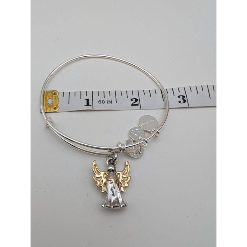Pode incluir: Uma pulseira de fio expans&iacute;vel em tom prateado com um pingente de anjo bicolor com asas em tom dourado e corpo prateado. Uma fita m&eacute;trica branca colocada atr&aacute;s da pulseira indica um di&acirc;metro de aproximadamente 6,4 cm.