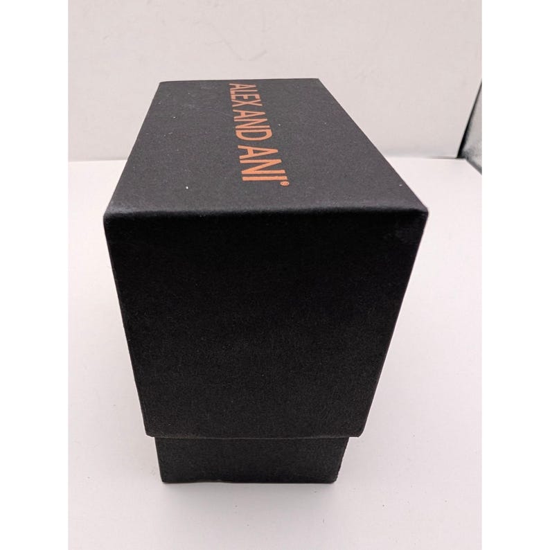 Pode incluir: Uma caixa de joias retangular preta com acabamento texturizado. A tampa superior apresenta o nome da marca ALEX AND ANI impresso em uma fonte de cor cobre met&aacute;lico. A caixa est&aacute; orientada verticalmente, mostrando claramente o topo e a frente.