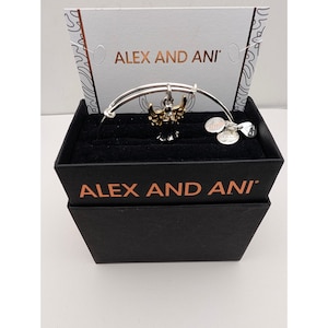 Pode incluir: Uma pulseira de fio expans&iacute;vel em tom prateado da Alex and Ani com um pingente central de anjo com asas em tom dourado e corpo preto. A pulseira inclui tr&ecirc;s pequenas etiquetas met&aacute;licas circulares e &eacute; apresentada numa caixa preta com cart&atilde;o.
