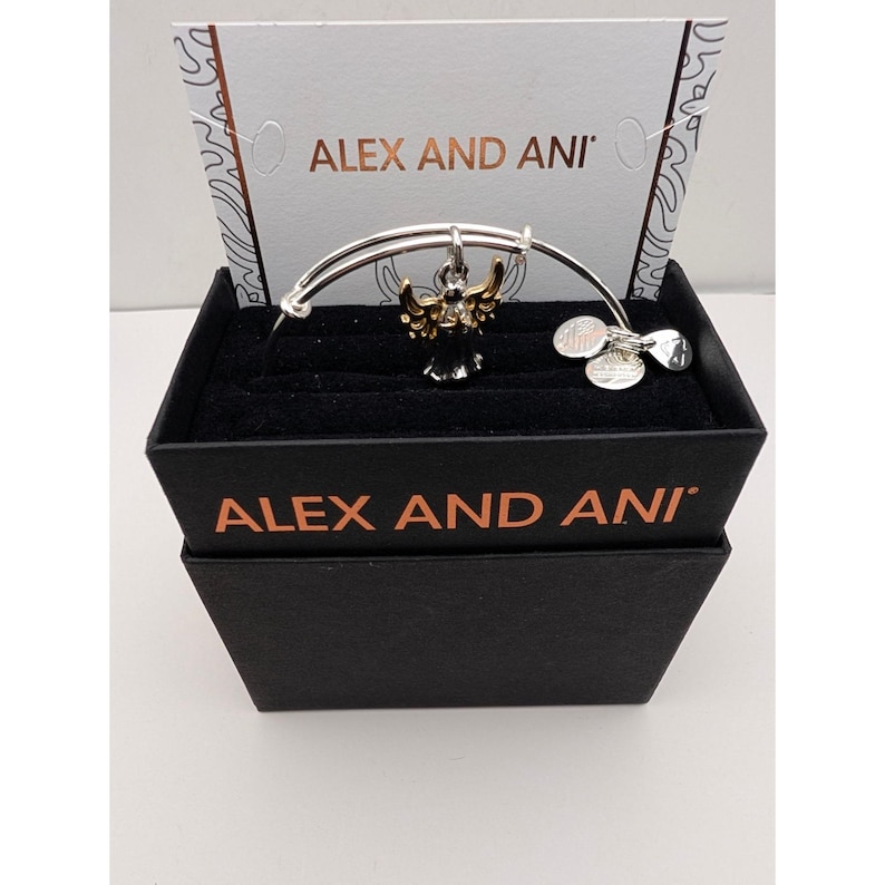 Pode incluir: Uma pulseira de fio expans&iacute;vel em tom prateado da Alex and Ani com um pingente central de anjo com asas em tom dourado e corpo preto. A pulseira inclui tr&ecirc;s pequenas etiquetas met&aacute;licas circulares e &eacute; apresentada numa caixa preta com cart&atilde;o.
