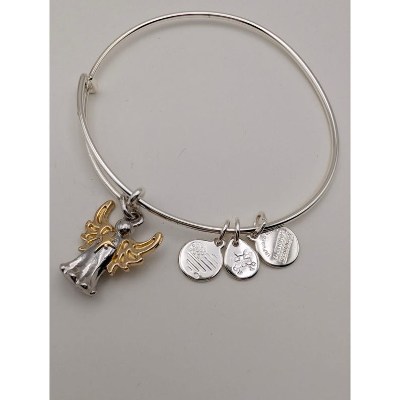 Pode incluir: Uma pulseira de fio expans&iacute;vel em tom prateado com um pingente de anjo bicolor com asas douradas e corpo prateado. Tr&ecirc;s pequenos pingentes circulares prateados adicionais est&atilde;o anexados, incluindo um com um design de cora&ccedil;&atilde;o e outro com texto.
