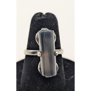 May include: A silver ring featuring a rectangular, gray stone. The stone is vertically oriented and set between two silver accents. The ring is displayed on a black ring holder.