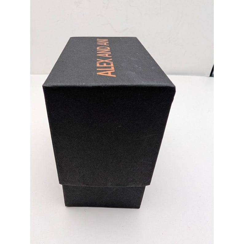 Pode incluir: Uma caixa retangular de papel&atilde;o preto com acabamento fosco. A superf&iacute;cie superior apresenta o nome da marca ALEX AND ANI impresso em uma fonte de cor cobre met&aacute;lico. A caixa est&aacute; orientada verticalmente, mostrando o topo e um lado.