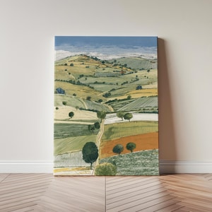 May include: A canvas print of a landscape featuring green hills, fields, and trees under a blue sky. A dirt track meanders through the fields. The artwork is propped against a wall.