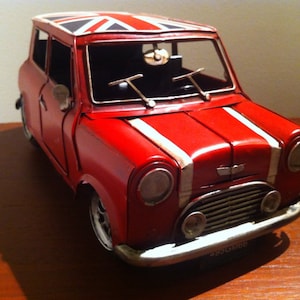 Pode incluir: Um carro de brinquedo vermelho com faixas brancas e um teto com a bandeira britânica. O veículo em miniatura tem faróis redondos, um para-choque cromado e uma placa de licença. O carro é um modelo clássico Mini Cooper.