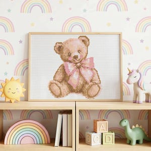 May include: A framed cross-stitch of a teddy bear with a pink bow. The bear is light brown with a pink bow. The background is white with a repeating pattern of rainbows and stars. The framed picture is on a wooden shelf with other toys.