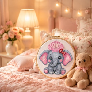 May include: A circular wooden embroidery hoop containing a cross-stitch design of a gray baby elephant with large pink ears, a pink seashell on its head, and a small red bow. The elephant has dark eyes and is depicted in a seated position.