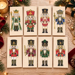 Könnte beinhalten: Acht rechteckige Stoffpaneele mit Kreuzstichmotiven von Nussknackerfiguren in verschiedenen Uniformen, Farben und Posen. Jedes Paneel zeigt eine eigene Figur, darunter Könige und Soldaten, die detailliert auf cremefarbenem Stoff gestickt sind.