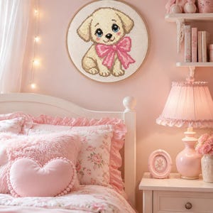 May include: A circular wooden embroidery hoop containing a cross-stitch design of a light tan puppy with large blue eyes and a bright pink bow around its neck. The puppy is depicted in a sitting position against a white fabric background.
