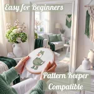 May include: A cross-stitch hoop featuring a cartoon goose wearing a green outfit and hat. The text "Easy for Beginners" and "Pattern Keeper Compatible" are visible. The scene is set in a bright room with a white sofa and green accents.