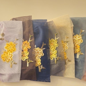 May include: A selection of sheer fabric pieces in a range of colours, each featuring embroidered yellow floral designs. The fabrics vary from cream to black, displaying a spectrum of shades and textures.