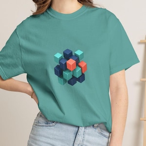 May include: A teal t-shirt featuring a geometric cube design. The design is composed of various colored cubes, including teal, navy blue, and coral. The shirt is made of a soft material and has short sleeves.