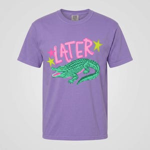 May include: A purple t-shirt featuring a green alligator graphic with the word "LATER" in pink, accented by yellow and pink stars. The alligator has a detailed, textured design. The shirt is a short-sleeved, crew neck style.