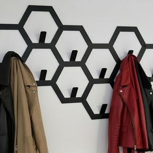 May include: Black honeycomb-shaped wall-mounted coat rack with multiple hooks. A black leather jacket, a tan jacket, and a red leather jacket hang from the hooks. The rack is mounted on a white wall.
