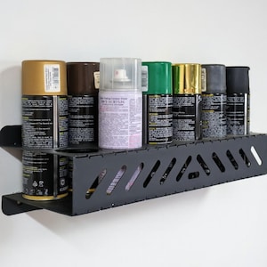 Puede incluir: Un organizador de pintura en aerosol de metal negro montado en una pared blanca, que contiene varias latas de pintura en aerosol en varios colores, incluyendo dorado, marrón, verde y negro. El organizador tiene un diseño ranurado.