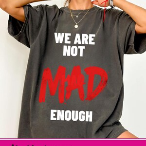 Puede incluir: Camiseta gris oscuro con el texto "WE ARE NOT MAD ENOUGH" en blanco y rojo. La camiseta tiene un corte holgado y está hecha de un material suave. La parte inferior de la imagen tiene el texto "digital PNG NO SVG INCLUDED".