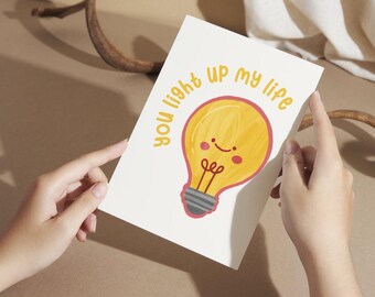 May include: A white greeting card with the text "you light up my life" and a cartoon lightbulb illustration. The lightbulb is yellow with a smiling face and red accents. The card is held by two hands.