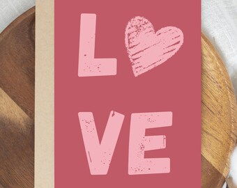 May include: A greeting card with the word "LOVE" in a distressed, light pink font on a red-pink background. A light pink heart is above the "VE". The card is set on a wooden tray.