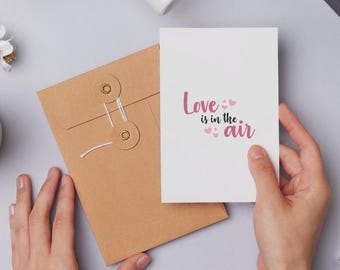 May include: A white greeting card with the text "Love is in the air" in pink, held by a person's hand. A brown envelope with a string closure is next to the card. The background is a light gray surface.