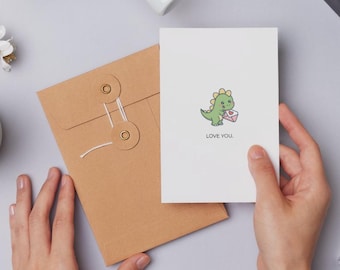 May include: A white greeting card with a cartoon dinosaur holding a heart-shaped envelope, with the words "LOVE YOU." A brown envelope with string closure is next to the card. The background is a light gray surface.