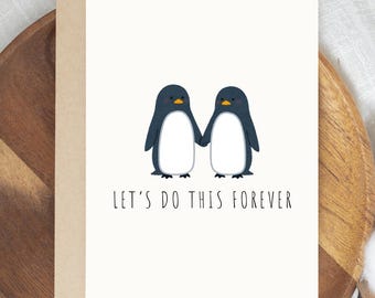 May include: A greeting card featuring two cartoon penguins holding hands. The penguins are black and white with yellow beaks. The text "LET'S DO THIS FOREVER" is printed in black below the penguins. The card is on a wooden tray.