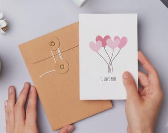 May include: A white greeting card with pink heart-shaped balloons and the text "I LOVE YOU". The card is held in a person's hands, next to a brown envelope with a string closure.