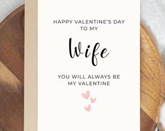 May include: A Valentine's Day card with the message "Happy Valentine's Day to my Wife. You will always be my Valentine." The card has a cream-colored front with black text and pink heart illustrations. The card is set against a wooden background.