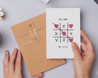 May include: A white greeting card with a tic-tac-toe game design, featuring hearts and the text "YOU WON MY HEART." The card is held by a person's hand, next to a brown envelope with a string closure.