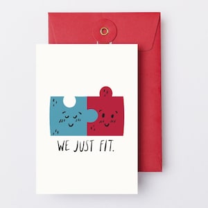 May include: A white greeting card with a blue and red puzzle piece design, with the text "WE JUST FIT." The card is paired with a red envelope.