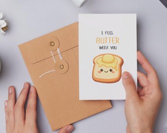 May include: A greeting card with the text "I FEEL BUTTER WITH YOU" and an illustration of a slice of toast with butter. The card is held in a person's hand, next to a brown envelope.