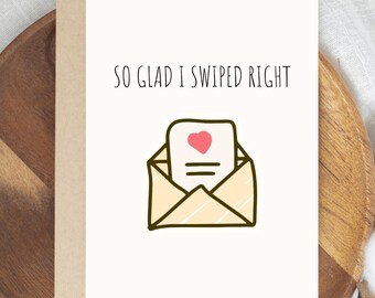 May include: A greeting card with the text "SO GLAD I SWIPED RIGHT" in black. The card features a cartoon illustration of an open envelope with a heart and lines on the letter inside. The card is on a wooden tray.