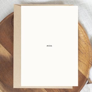 Pode incluir: Um cartão de felicitações em branco com a palavra "mine" em uma fonte pequena e preta. O cartão é branco sujo e está colocado sobre um envelope marrom claro. O cartão e o envelope estão em um prato de madeira.