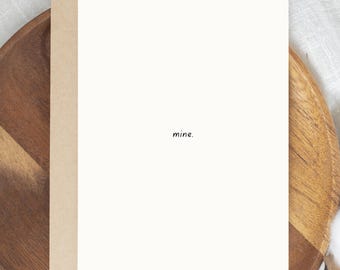 May include: A blank greeting card with the word "mine" in a small, black font. The card is off-white and set against a light brown envelope. The card and envelope are on a wooden plate.