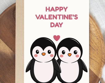 May include: Valentine's Day greeting card featuring two cartoon penguins with black and white bodies, orange feet, and rosy cheeks. The words "HAPPY VALENTINE'S DAY" are in pink above the penguins, with a pink heart between them. The card is on a wooden tray.