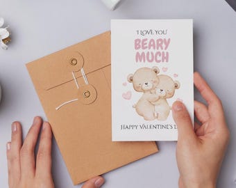 May include: A Valentine's Day card with two hugging teddy bears and the text "I Love You Beary Much". The card is held near a brown envelope with a string closure. Pink hearts are scattered around the bears.